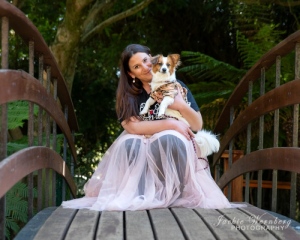 woman-wearing-pink-frilly-dress-knealing-on-bridge-holding-small-fluffy-dog