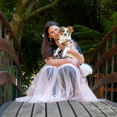 woman-wearing-pink-frilly-dress-knealing-on-bridge-holding-small-fluffy-dog
